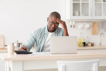 Stressed mature black man using calculator and laptop computer, calculating taxes, checking monthly expenses