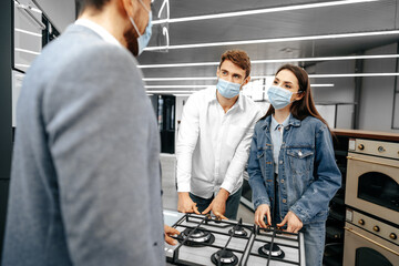 Shop assistant in hypermarket shows new model of gas stove to young couple, all wearing medical...