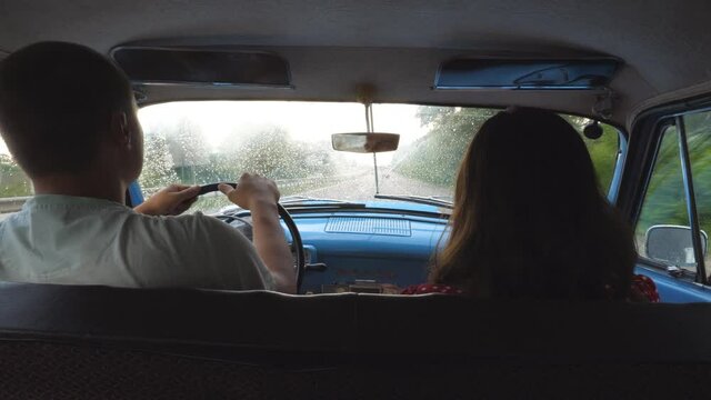 Young couple riding at vintage car during rainy wet weather. Unrecognizable man and woman sitting at the front seat of old retro car at road trip. Travel concept. Slow motion Rear back view