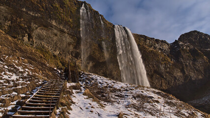 Stunning low angle view of famous waterfall Seljalandsfoss in on the southern coast of Iceland near ring road on sunny winter day with wooden stairs and snow-covered brown grass.