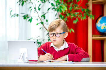 Smart boy in glasses using tablet computer. Child writing homework or preparing for the exam. Teen using laptop to study. Student. School. Remote. Online learning. New normal. Home Based Learning.