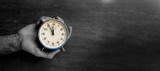 In the man's hand is an old analog mechanical retro clock. Below is a wooden brown background. The time on the clock is five minutes to twelve o'clock.