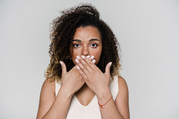 The portrait of a curly-haired woman looking at the camera and covering her mouth with hands in the studio
