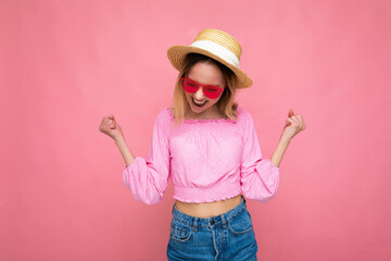 Photo shot of attractive happy smiling young blonde woman wearing summer casual clothes and stylish sunglasses isolated over colorful background wall and showing yeah gesture