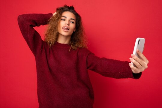 Sexy Beautiful young woman with curly hair wearing dark red sweater isolated on red background wall holding and using smart phone looking at telephone screen and taking selfie and showing tongue