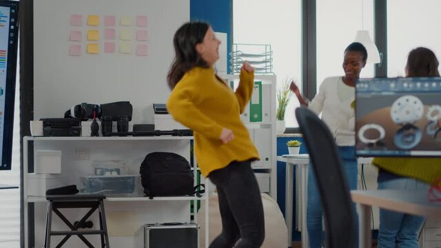 Happy diverse businesswomen dancing in company office celebrating success of project, singing and having fun during working hours. Excited multiethnic engineers using computer showing cad software