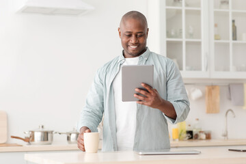 Positive african american mature man reading news on digital tablet and drinking coffee in kitchen