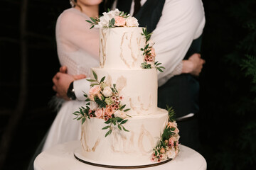Luxury wedding tiered white cake decorated with flowers, photo at night on the background of the bride and groom