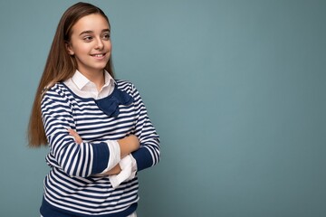 Shot of beautiful positive smiling brunette little girl wearing striped longsleeve standing isolated over blue background wall crossing hands and looking to the side