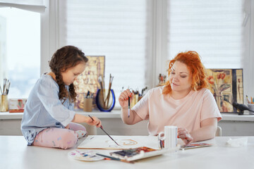 Obraz premium Art creative leisure activity. Beautiful caring art teacher or painter helps to draw cute preschool girl sitting at the table using watercolor, teacher enjoy helping small kid doing painting at home