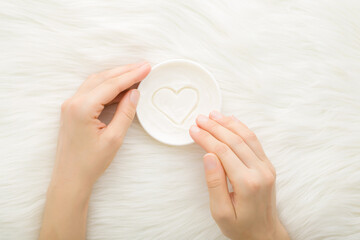 Young adult woman hands using white cream jar with heart shape on light fur blanket background. Care about clean and soft body skin. Closeup. Point of view shot. Top down view.