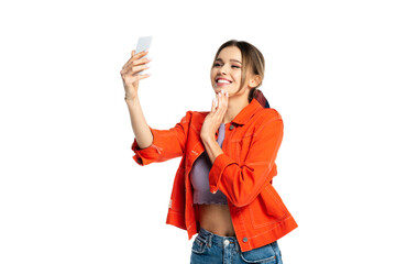 Naklejka premium cheerful young woman in crop top and orange shirt taking selfie on smartphone isolated on white