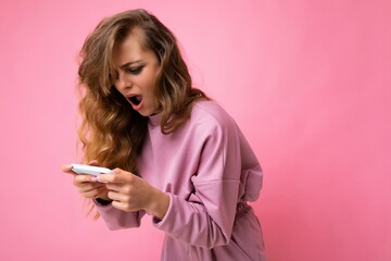 Side-profile amazed surprised blonde young woman with open mouth wearing pink hoodie isolated over pink background with copy space holding and playing games via telephone looking at device display