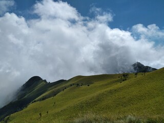 A panoramic view of rolling green hills with fluffy clouds covering the mountain peaks. The image captures the serene beauty of the natural landscape.
