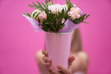 A little girl in pink dress with bouquet of mixed flowers in a paper cup, hid behind the flowers. The girl holds out her hands and gives flowers.