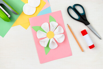 A flower made of cotton pads. Colored paper, stapler, scissors, scissors, pencil on a white table. Children's craft card for mother's day. Step-by-step instruction. Step 12.