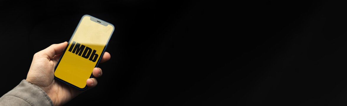 Kharkov, Ukraine - April 20, 2021: iMDB app logo banner with black background, copy space, man uses smartphone with application