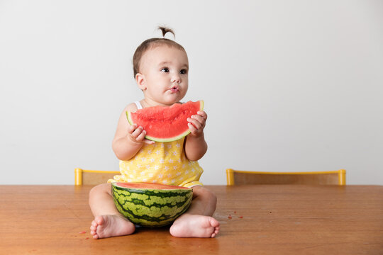 Cute baby girl sitting on kitchen table holding slice of watermelon