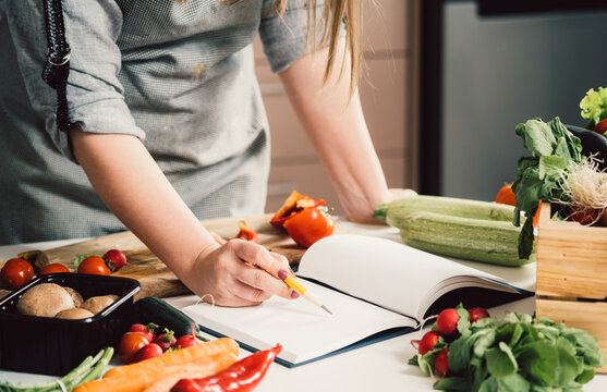 Unrecognizable woman is looking for recipes in cookbook.
Female chef reading recipes in book and holding pen on table full with fresh vegetables for healthy lunch