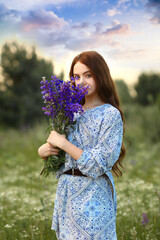 A beautiful young redhead girl with blue eyes holds a bouquet of wildflowers in her hands. Standing in nature at sunset time in a meadow where there are many wildflowers. Summer. Nature concept.