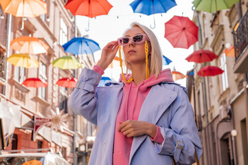 Obraz na płótnie Canvas Fashionable woman wearing trendy pink sunglasses with yellow plastic chain, hoodie, blue trench coat, walking in European city. Streetstyle, street fashion conception. Copy, empty space for text