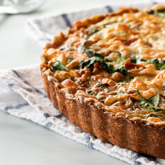 Close up of a homemade baked spinach and bacon quiche on a kitchen towel cooling by a bright window. Instagram crop.