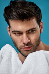 Fototapeta premium Studio portrait of sexy brunette guy with wet washed face holding white towel, looking away, posing isolated over blue background
