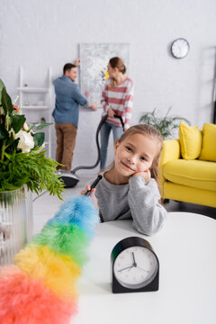 Smiling Kid With Dust Brush Looking At Camera Near Clock And Plants On Table.