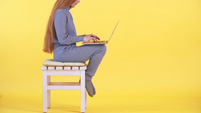 A woman with long ginger hair is sitting in casual costume and socks on the chair and working online on the laptop. Yellow background