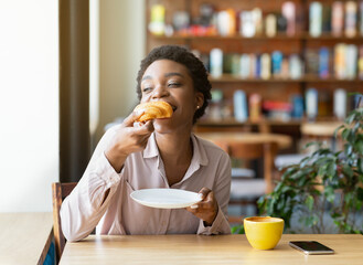 Pretty black woman enjoying breakfast at cafe, eating fresh croissant and drinking aromatic coffee