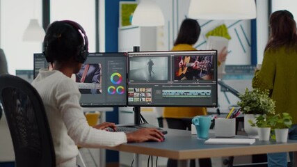 Black videographer putting on headset editing movie using post production software working in creative start up agency office. African editor processing video footage on computer with dual displays