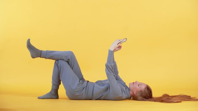Young woman with freckles and long ginger hair is lying on the floor and making selfie on her smartphone. Yellow background