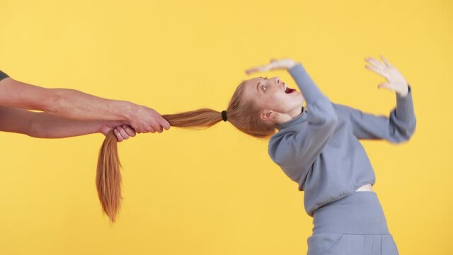 Male hands are pulling girl's ponytail. Young woman with freckles is surprised and scared. Yellow background
