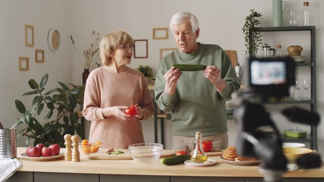 Positive senior man and woman showing ingredients and speaking on camera while filming culinary vlog together at home