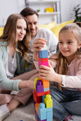 Obraz premium Smiling girl on blurred background playing building blocks near parents.