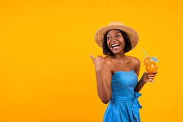 Happy black woman in summer dress and hat drinking refreshing cocktail, pointing at empty space on orange background
