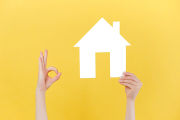Fototapeta na wymiar Female hands holding small paper white house model and makes okay gesture, isolated over yellow studio background wall with copy space for advertisement. Acceptance okay and mortgage concept