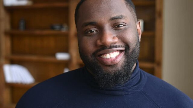 Close-up cropped video portrait of handsome African-American guy, bearded joyful black guy looking at the camera and smiles wide and toothy, attractive dark-skinned male in turtleneck laughs indoor