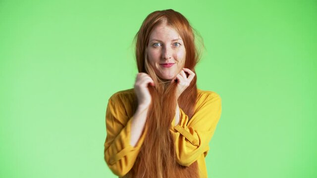 Woman is having her long ginger hair on face, then looking through them into the camera, smiling and waving hand. Green background