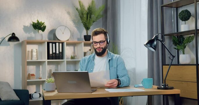 Remote business concept where attractive smiling successful young bearded guy in headset sitting in front of computer during video chat with coworker