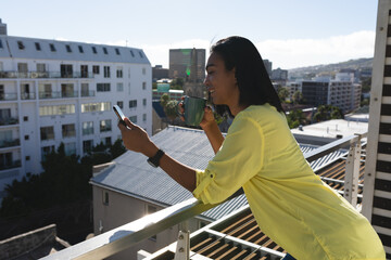 Smiling mixed transgender woman man standing on sunny roof terrace holding coffee using smartphone