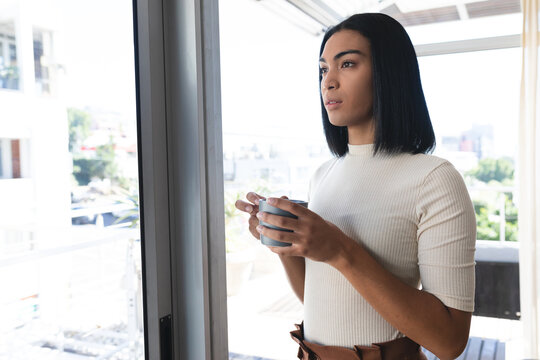 Mixed race transgender woman standing looking out of window holding cup of coffee