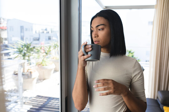 Mixed race transgender woman standing looking out of window holding cup of coffee