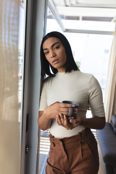 Portrait of mixed race transgender woman standing by window holding cup of coffee