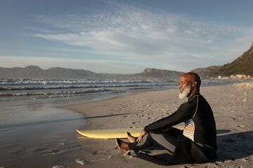 African american senior man on beach sitting with surfboard looking out to sea