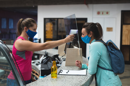 Female receptionist and customer wearing masks checking temperature over the counter at gym
