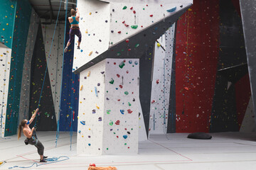 Two caucasian women wearing face masks using ropes to climb wall at indoor climbing gym