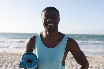 Portrait of smiling african american man exercising outdoors, holding yoga mat on beach