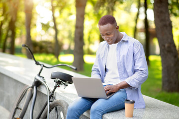 Distance work. Joyful African American man with bicycle working on laptop computer at urban park, empty space