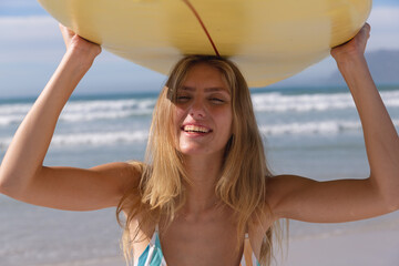 Smiling caucasian woman wearing bikini carrying yellow surfboard on her head at the beach